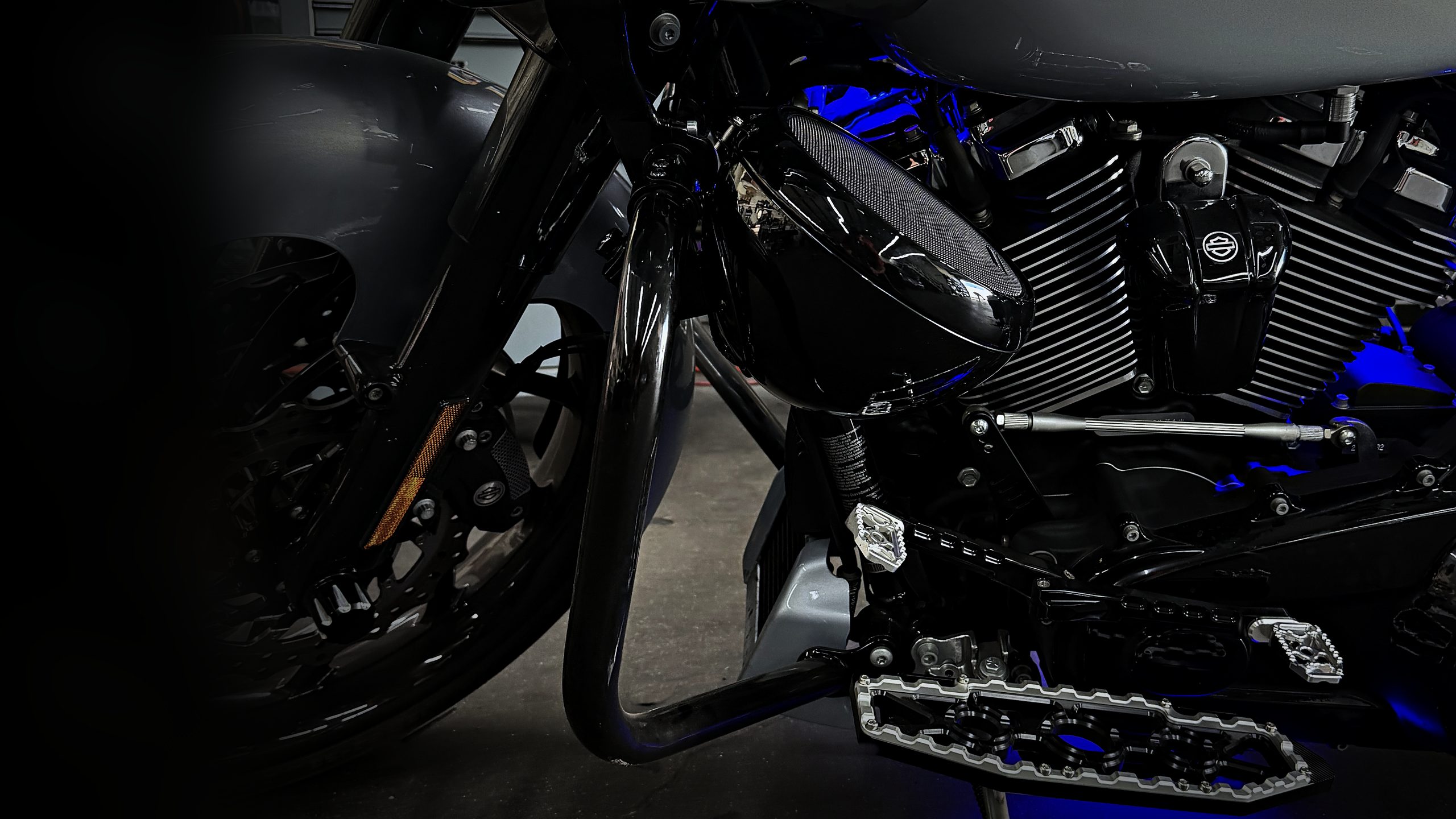 Black AMS speaker pod mounted on a Harley crash bar with its mounting bracket, lit by blue LED, at Audio MotorSports in Carson